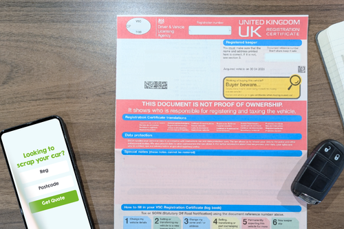 Photo of a V5C registration document, a phone showing a scrap car quote form and a set of car keys all on a wooden table top