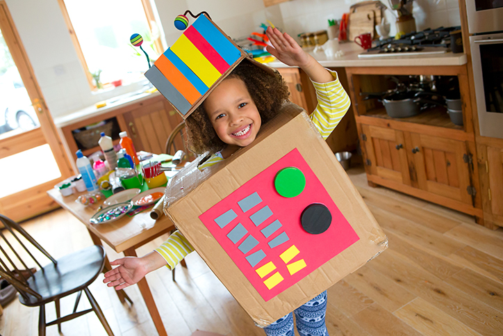 Little girl is standing in her kitchen, pretending to be a robot. She has made a body and hat using cardboard boxes. The arts and crafts are spread across the dining table in the background. She is smiling at the camera and has her arms positioned like they are stiff, mimicking a robot.