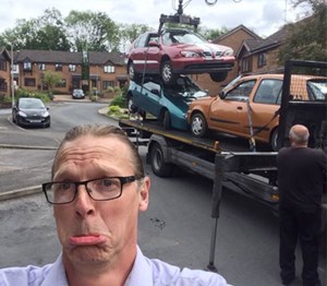 Man with glasses looking sad, in the background is a truck with old cars loaded on